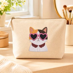 May include: A beige canvas zippered pouch featuring an embroidered calico cat wearing pink heart-shaped sunglasses and a dark brown scalloped collar. The cat has white, orange, and black fur patches, and the pouch has a metallic zipper closure.