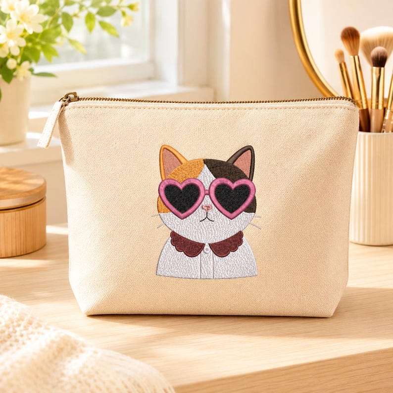 May include: A beige canvas zippered pouch featuring an embroidered calico cat wearing pink heart-shaped sunglasses and a dark brown scalloped collar. The cat has white, orange, and black fur patches, and the pouch has a metallic zipper closure.