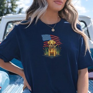 May include: Navy blue t-shirt featuring a golden retriever holding a duck in its mouth, set against an American flag backdrop. The dog is standing in water with reeds. A patriotic design for dog lovers.