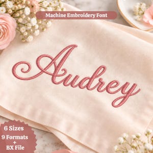 Könnte beinhalten: Ein hellcremefarbener Stoff mit dem Namen Audrey, der in einer altrosa Schreibschrift gestickt ist. Der Text Machine Embroidery Font steht oben, und eine sternförmige Grafik unten zeigt 6 Sizes, 9 Formats + BX File.