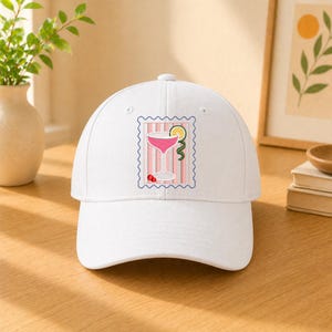 May include: A white baseball cap featuring a centered embroidered graphic of a pink cocktail in a stemmed glass. The design is framed by a blue scalloped border on a background of vertical pink and white stripes, with a lime slice and red cherry.