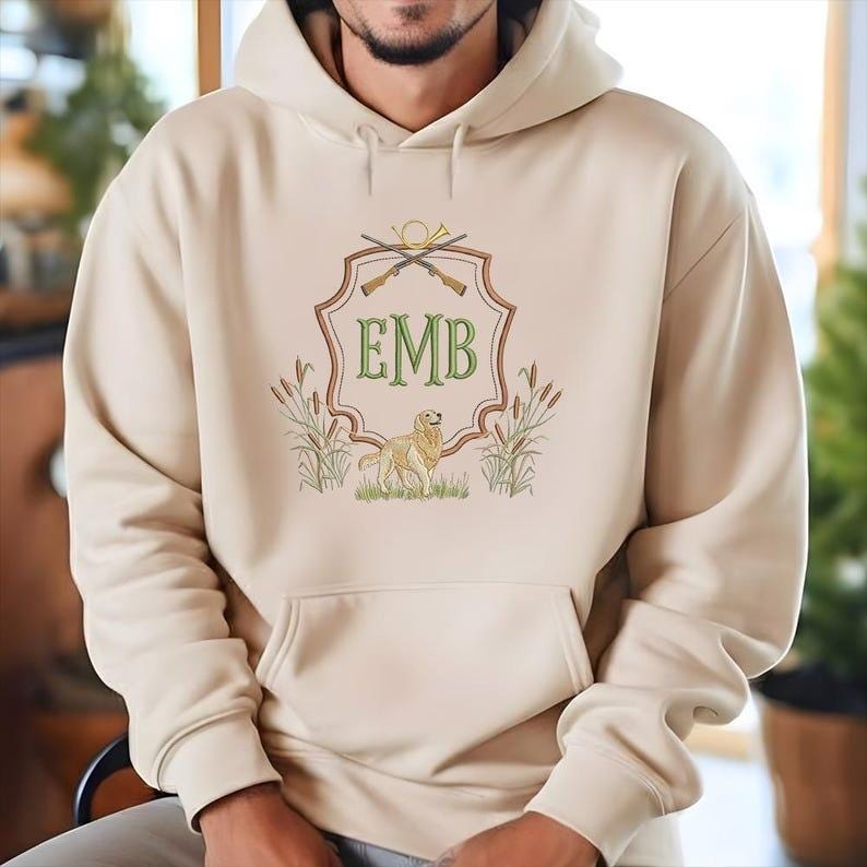 May include: Beige hoodie with a front pocket and a hood. The front features an embroidered crest with crossed rifles, a hunting horn, and the initials "EMB" in green. A golden retriever dog is also embroidered on the crest.
