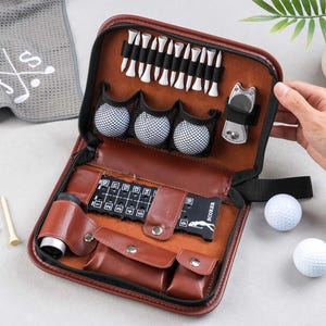 Peut inclure: Un kit de golf en cuir marron avec divers accessoires, dont des balles de golf, des tees, un marqueur de score et un outil de réparation de divot. Le kit est ouvert, révélant le contenu, et est accompagné d'une serviette de golf grise.