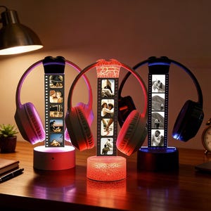 Puede incluir: Tres soportes iluminados para auriculares con tiras de fotos personalizadas. Los soportes cuentan con iluminación roja, morada y azul, con auriculares a juego. Los soportes están sobre un escritorio de madera con una lámpara, un cuaderno y un reloj.
