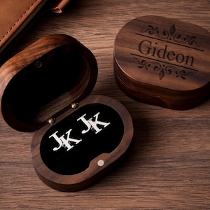 May include: Oval wooden jewelry box with a hinged lid, open to reveal a black interior with silver initials "JK JK". The closed box has the name "Gideon" engraved on the lid. The box is made of dark wood and sits on a wooden surface.