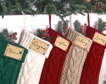 Op de afbeelding: Vijf kabelgebreide kerstsokken in groen, crème, rood en beige. Elke sok heeft een gepersonaliseerd naamlabel en is versierd met nep groen en bessen. De sokken hangen aan een wit oppervlak.
