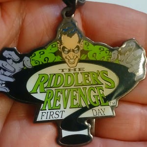 May include: A metal keychain featuring the Riddler from Batman. The keychain is shaped like a question mark and features the text "The Riddler's Revenge - First Day" with a green and black color scheme.