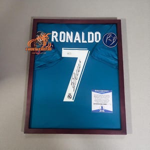 May include: Framed teal soccer jersey with the name "RONALDO" and the number "7" in white. The jersey is signed and includes a Beckett Certificate. The frame is a dark brown color.