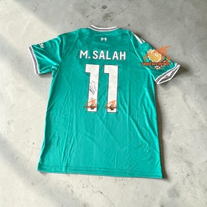 May include: A teal soccer jersey with the name "M. SALAH" and the number "11" printed on the back. The jersey has white and black trim on the sleeves and collar. The jersey also has a signature on the number 11.