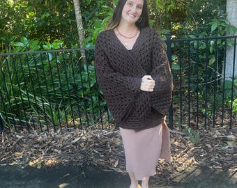 May include: A dark brown, oversized crochet sweater with wide sleeves. The sweater is paired with a light pink dress and white sandals. The background features a black fence and lush greenery, creating a casual, outdoor aesthetic.