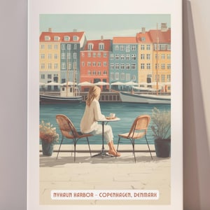 May include: A framed print of Nyhavn Harbour, Copenhagen, Denmark. The image shows a person at a table with colourful buildings in the background. Two chairs and potted plants are either side of the table. The print is displayed on a white surface.