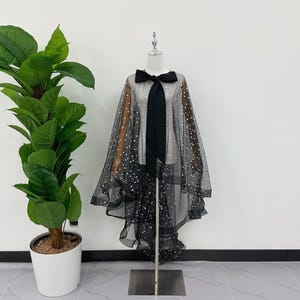 May include: A black sheer cape with a bow at the neck and a long black tie. The cape is decorated with silver glitter and has a ruffled hem. The cape is displayed on a mannequin.