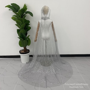 May include: A sheer, grey bridal cape with a hood and sparkling embellishments. The cape is displayed on a mannequin and has a long train. The words "PrisonerUnitedStates Handmade Veils" are in the bottom right corner.