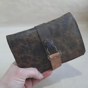 Könnte beinhalten: Eine braune Ledergeldbörse mit Used-Look. Die Geldbörse hat einen dunkelbraunen Riemen mit einem dekorativen Knopf und einen helleren braunen Wildlederakzent. Die Geldbörse wird in einer Hand gehalten.