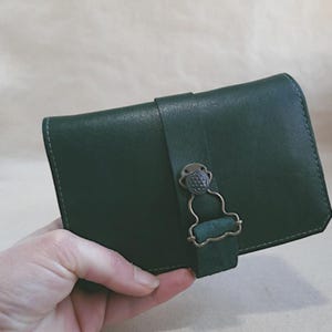 May include: A dark green leather wallet with a decorative clasp. The wallet is rectangular with a flap that secures with a strap and a metal clasp. The leather appears to be smooth and the stitching is visible along the edges.