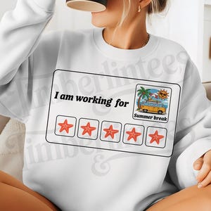 Op de afbeelding: Witte sweatshirt met de tekst "I am working for" boven een afbeelding van vijf zeesterren en een strandscène met een busje met de tekst "Summer break". Een persoon houdt een zwarte mok vast.