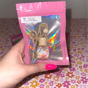 May include: A clear, faceted glass car air freshener with a wooden top and a woven cord, packaged in a pink holographic bag. The label reads "Watermelon Lemonade @CreationsByLeo".