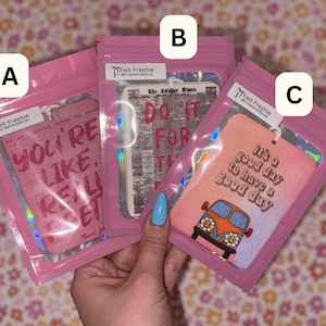 May include: Three air fresheners in pink packaging. One has the text "YOU'RE LIKE, REALLY, REALLY, GET IT". Another says "DO IT FOR THE DAY". The third says "It's a good day to have a good day".