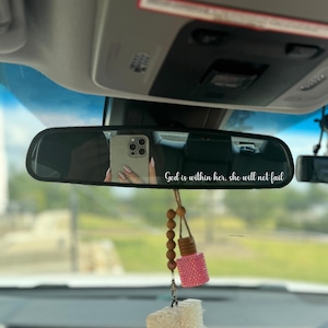 May include: A car's rearview mirror reflects a person holding a phone. The mirror has the text "God is within her, she will not fail." A pink, bedazzled air freshener hangs below the mirror, along with a string of wooden beads.