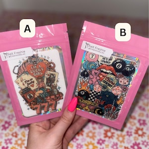 May include: Two pink pouches containing air fresheners. One features a tiger, playing cards, and the text "I'M A LUCKY GIRL". The other has a cowboy theme with dice, flames, and the text "HE'S A COWBOY".