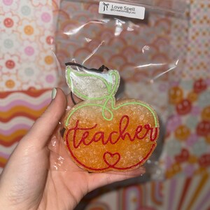 May include: Orange apple-shaped air freshener with the word "teacher" in red script and a heart. The apple has a green stem and leaf design. The air freshener is in a clear plastic bag with a black string for hanging.