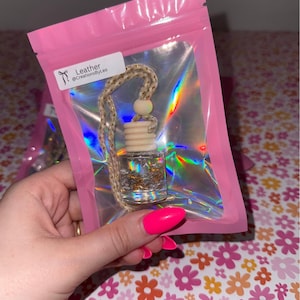 May include: A clear glass car air freshener with gold flakes and a wooden top, hanging from a braided cord. The freshener is in a pink plastic bag with a label that says "Leather @CreationsbyLeo".