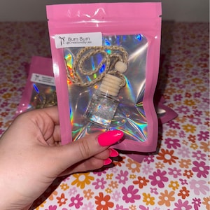 May include: A clear glass car air freshener with a wooden cap and a decorative rope. The air freshener is inside a pink holographic bag with the text "Bum Bum @CreationsbyLeo". The background is a floral pattern.