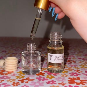 May include: A clear glass bottle with a gold dropper is held above a small, empty glass bottle and a bottle with a label that reads "Bum Bu @Creations". The bottles sit on a floral patterned surface.