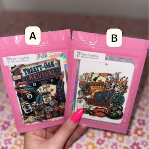 May include: Two air fresheners in pink packaging. One features a design with the text "Treaty Oak Revival", lips, and other graphics. The other has a similar design with the same text and additional graphics. Both are labeled "Felt Freshie".