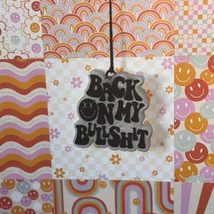 May include: A clear, glittery charm with the phrase "BACK ON MY BULLSH*T" in black, hanging from a black string. The charm features a smiley face with crossed-out eyes. The background is a collage of patterned papers with rainbows, flowers, and smiley faces.