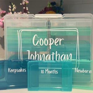 May include: Clear plastic keepsake storage containers with teal and blue accents. The containers are labeled "Cooper Johnathan", "Keepsakes", "10 Months", and "Newborn". The containers are designed to store baby keepsakes.