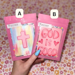 May include: Two pink air fresheners in clear plastic bags. One has a pink cross on a colorful background. The other says "GOD IS GOOD" with hearts. Both have a small white tag with text.