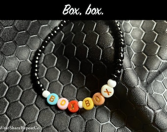 May include: A beaded bracelet spelling "BOX BOX" with colourful letter beads. The bracelet has black beads and is set against a textured black background. The text "Box, box." is at the top.