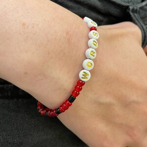 May include: A white beaded bracelet with the word "WORDS" spelled out in gold letters. The bracelet is made with red and black beads.