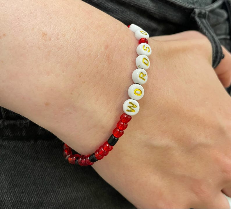 May include: A white beaded bracelet with the word "WORDS" spelled out in gold letters. The bracelet is made with red and black beads.
