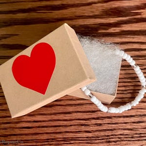 May include: A white beaded bracelet with silver accents in a small brown box with a red heart on the lid.