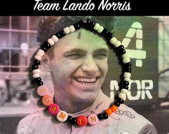 May include: A black and white beaded bracelet with the letters "TEAM LN" spelled out in colourful beads. The bracelet is worn by a person with a smiling face. The background is a blurry image of a racing car.