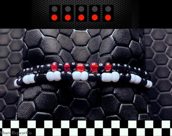 May include: A black and white beaded bracelet with red accents. The bracelet is wrapped around a textured black surface. The top of the image features a series of red lights. The bottom of the image has a black and white checkered pattern.