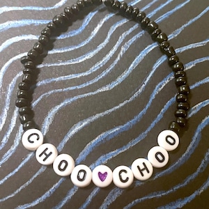 May include: A black beaded bracelet with white letter beads spelling "CHOOCHOO" and a purple heart bead. The bracelet is on a dark background with blue and white wavy lines.