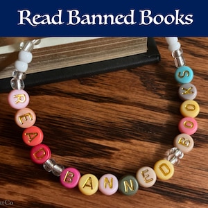 Puede incluir: Un brazalete de cuentas de colores con las palabras "Read Banned Books" escritas en letras doradas. El brazalete está sobre una superficie de madera con un libro en el fondo.