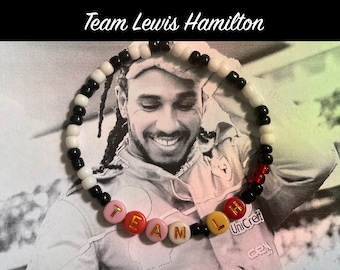 May include: Black and white beaded bracelet with red beads and white letter beads spelling "TEAM LH". The bracelet is worn by a person in a racing suit with the text "UniCredit" visible.
