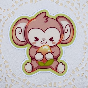 May include: A sticker featuring a cartoon monkey holding a fruit, set on a white, lace-edged doily. The monkey is light brown with a green outline, and the fruit is yellow and orange. The doily has a scalloped edge.