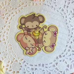 May include: A whimsical arrangement featuring a monkey sticker with bananas, surrounded by star-shaped and heart-shaped candies, chocolate pieces, and a croissant. A white doily and lined paper provide the backdrop, with a skewer of dango and floral accents.