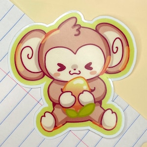 May include: A sticker featuring a cartoon monkey in shades of brown, holding a fruit. The monkey has large ears and a smiling face. The sticker is outlined in green and sits on lined paper.