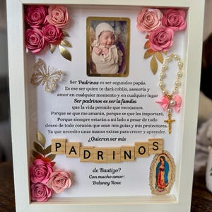 May include: A white picture frame with a pink and gold floral design. The frame contains a photo of a baby, a string of pearls, a gold cross, and a small picture of the Virgin Mary. The frame also includes the text "Ser Padrinos es ser segundos papás. Es ese ser quien te dará cobijo, asesoría y amor en cualquier momento y en cualquier lugar Ser padrinos es ser la familia que la vida permite que escojamos. Porque se que me amarán, porque se que les importaré, Porque siempre estarán a mi lado a pesar de todo deseo de todo corazón que sean mis guías y mis protectores. Ya que necesito unas manos extras para crecer y aprender. ¿Quieren ser mis PADRINOS de Bautizo? Con mucho amor: Delaney Rose"
