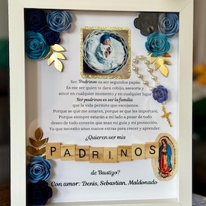 May include: A framed baptism keepsake with a baby photo, blue paper flowers, and gold accents. The text reads "Padrinos" and "¿Quieren ser mis de Bautizo?" in Spanish, with a Virgin Mary image and a rosary.