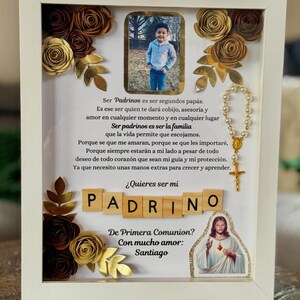 May include: A white framed shadow box with a photo of a child, paper flowers, and a rosary. The text reads "¿Quieres ser mi Padrino?" and "De Primera Comunión? Con mucho amor: Santiago."