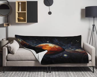 Op de afbeelding: Een pluche deken met een zwarte achtergrond en een kleurrijke galaxyscène met een grote oranje planeet. De deken is over een grijze bank gelegd met een wit kussen.