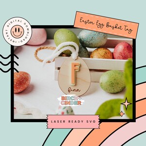 Op de afbeelding: Houten paasmandje tag met de letter "F" in perzik. De tag is bevestigd aan een wit touwtje. De tag staat voor een witte houten kist gevuld met kleurrijke paaseieren. De tekst "Easter Egg Basket Tag" staat bovenaan.