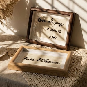 May include: Two framed artworks with handwritten text. The top frame is dark brown, the bottom frame is light brown. The top frame reads "Love always mama xxx". The bottom frame reads "love William". A vase with dried flowers and a teacup are also visible.